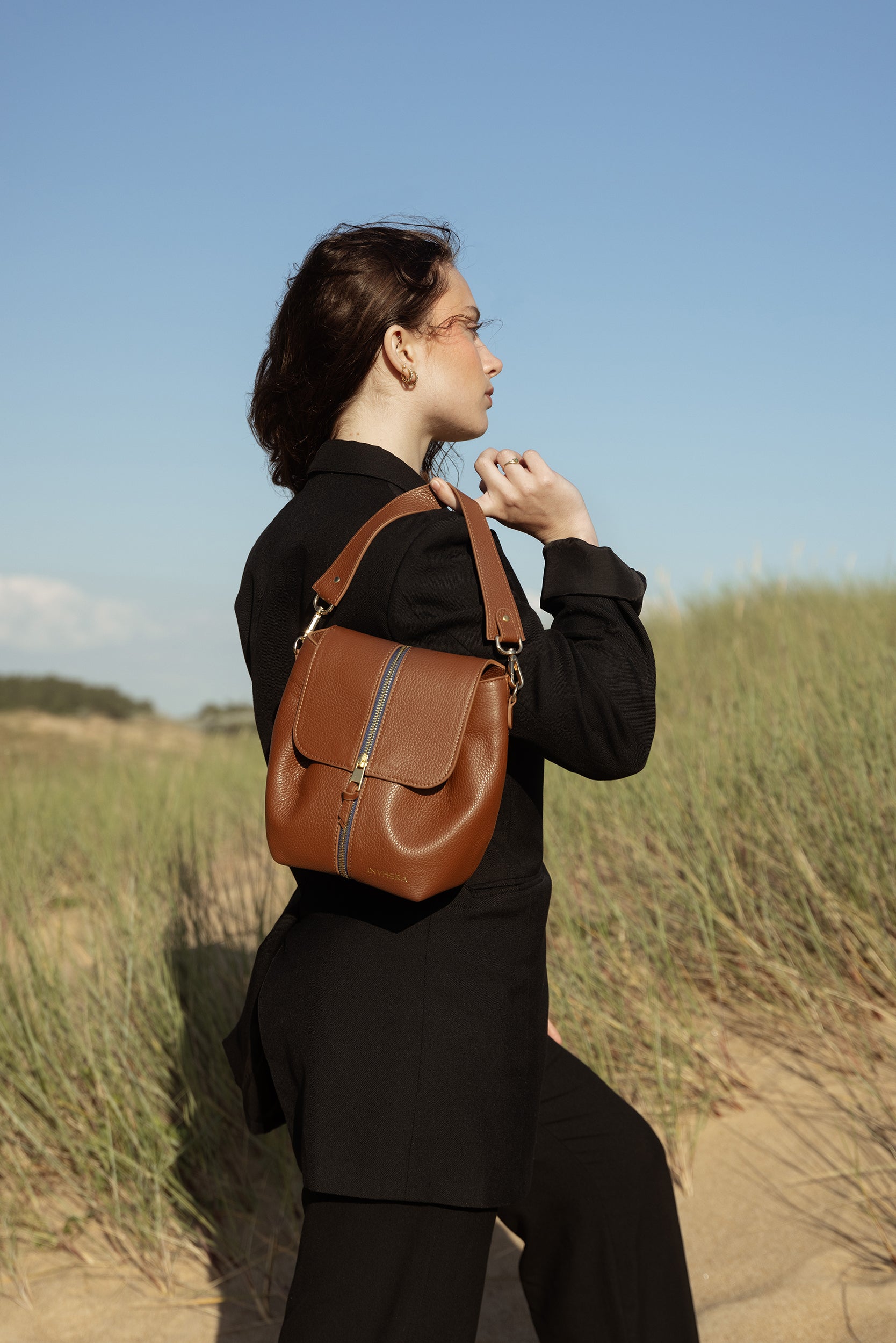 Une femme sur la plage habillée d'un ensemble blazer noir. Portant un sac à main de chez INVHERA, il est posé au dos de son épaule droite. Le sac à main est de couleur marron en cuir de vachette avec des jolies plis.