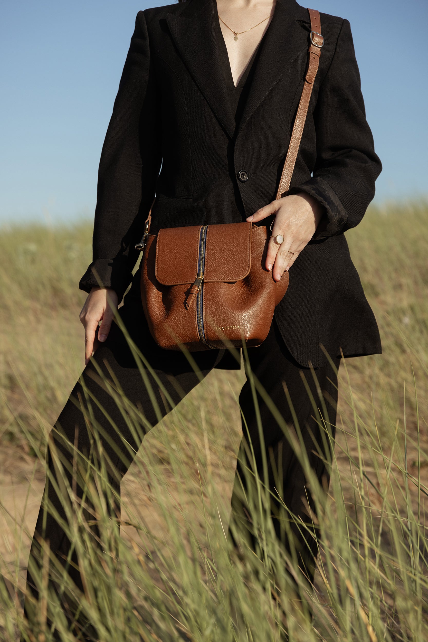 Une femme d’un ensemble élégant est debout, portant un sac à main à bandoulière de chez INVHERA ; Féminin et pratique en cuir de vachette grainé de couleur marron avec fermeture centrale. Plus qu’un sac bandoulière mais un sac à main unique.
