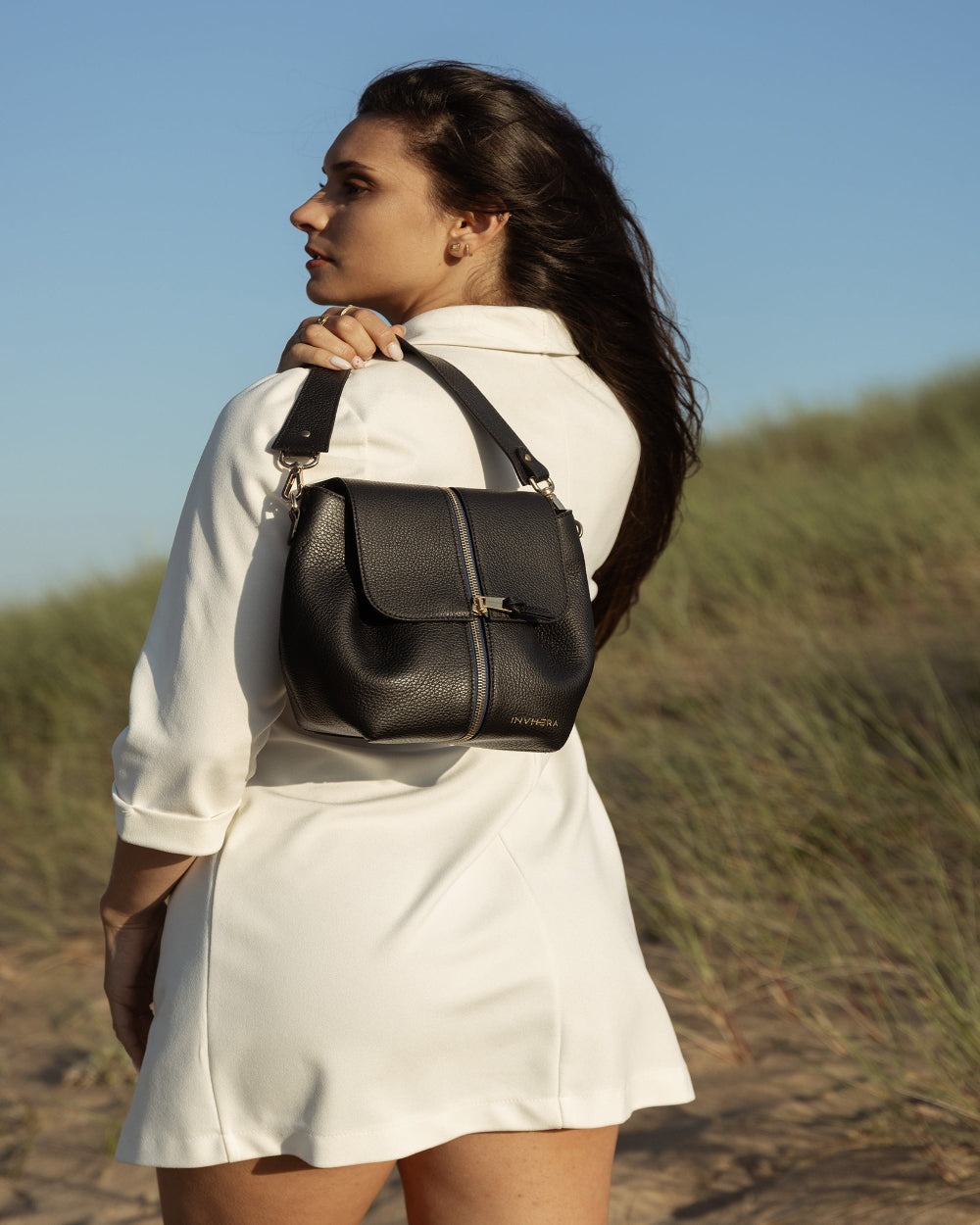 Une jeune femme moderne à la plage, portant un sac à main avec anse amovible de chez INVHERA en cuir de vachette grainé de couleur noir. Trouvez l’inspiration pour vos tenues du quotidien pour une allure élégante et audacieuse.